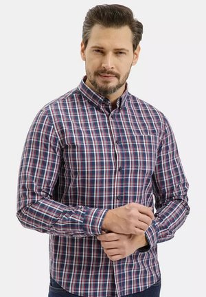 Mężczyzna o krótkich włosach i brodzie, noszący niebiesko-czerwono-białą koszulę w kratę zapinaną na guziki, poprawiający mankiet na neutralnym tle.