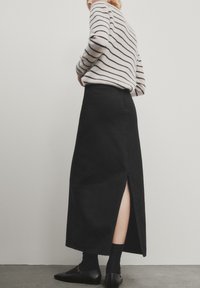 Jupe en denim noire, longueur cheville, avec une fente à l'arrière, associée à un pull en tricot à rayures et des chaussures noires à enfiler, présentée sur un fond neutre.