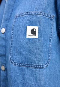 Denim shirt in light blue with a chest pocket, featuring a white square label with a black logo. Constructed from sturdy fabric with stitched details.