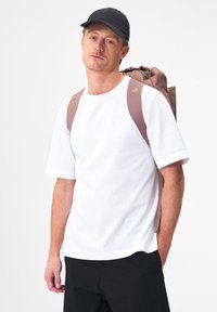 Weißes Baumwoll-T-Shirt, schwarze Mütze und stylischer brauner Rucksack mit geschnürtem Oberteil. Verfügt über gepolsterte Träger und abgerundete Kanten. Schlichter Hintergrund.