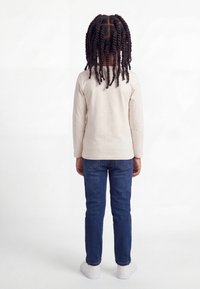 Παιδί με σκούρα, σγουρά μαλλιά στέκεται με την πλάτη στραμμένη, φορώντας μπεζ πουκάμισο με μακριά μανίκια, μπλε τζιν και λευκά αθλητικά παπούτσια σε λευκό φόντο.