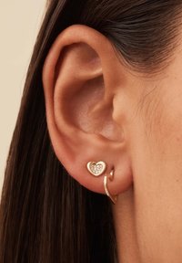 Des boucles d'oreilles en tone doré présentent un clou en forme de cœur avec des accents de pierres claires et un design de boucle spiralée. Des finitions en métal texturé rehaussent les détails.