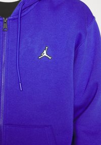 Gros plan d'un sweat à capuche bleu zippé avec une poche avant et un petit logo blanc en forme de silhouette d'une personne qui saute.