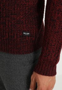 Lähikuva maroonin- ja mustan värin neulotun paidan hihasta ja helmasta, jolle on ommeltu "ONLY & SONS" -tagi. Asusteena harmaateksuroitu housu ja näkyvä käsi.