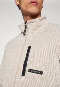 Μπεζ υφασμάτινο μπουφάν fleece με ψηλό γιακά, που διαθέτει μαύρη τσέπη στο στήθος με φερμουάρ και ταμπελάκι με το λογότυπο. Τα φερμουάρ έχουν αντίθετο χρώμα.