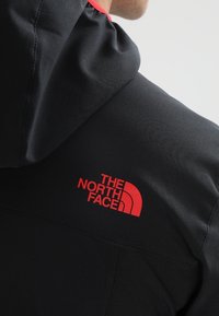 Personne portant une veste à capuche noire avec un logo rouge "The North Face" sur l'épaule, sur un fond gris clair.