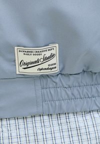 Tissu bleu clair avec une étiquette rectangulaire blanche cousue dessus indiquant « Jack & Jones Creative Dept. Originals Studio Copenhagen ©1990 ».