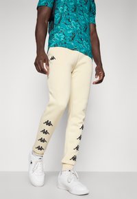 Beige joggingbroek met zwarte Kappa-logo's langs de zijkanten, gecombineerd met een groen abstract patroon shirt en witte sneakers.