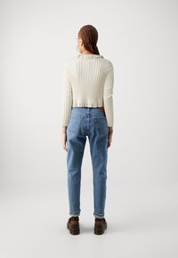 Žena s copánky, oblečená v krémovém žebrovaném svetru, modrých džínách, hnědých botách a s velkými kruhovými náušnicemi, stojící zády k fotoaparátu na bílém pozadí.