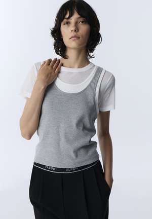 Woman with short dark hair wearing a white t-shirt under a gray tank top and black pants with "FLEUR" waistband, standing with one hand on her shoulder.