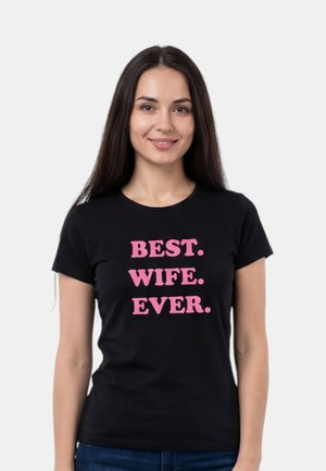 Junge Frau mit langen dunklen Haaren trägt ein schwarzes T-Shirt mit pinker Aufschrift "BESTE. FRAU. ÜBERHAUPT." vor schlichtem hellem Hintergrund.