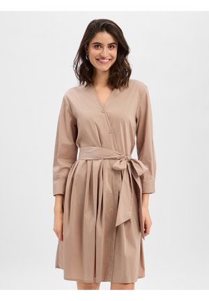 Uśmiechnięta kobieta o falowanych ciemnych włosach, ubrana w beżową, długorękawową sukienkę wiązaną w talii, stojąca na tle jasnej, jednolitej ściany.