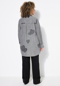 Camicia grigia a maniche lunghe con una sottile trama a righe, decorata con piccoli cuori neri. Abbinata a pantaloni neri a gamba larga, mostrata di spalle.