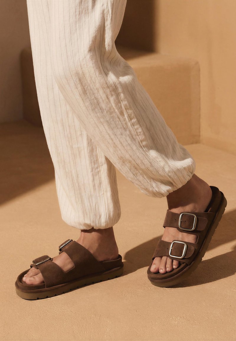 Persoon die losse, lichtgekleurde broek met fijne strepen en bruine suède sandalen met twee zilveren gespen draagt, staand op een beige ondergrond.