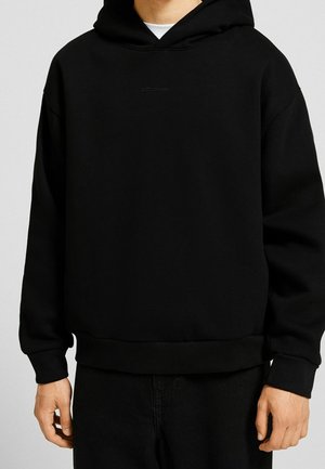 Mann trägt einen schlichten schwarzen Kapuzenpullover mit minimalem Text auf der Brust, kombiniert mit schwarzen Hosen.