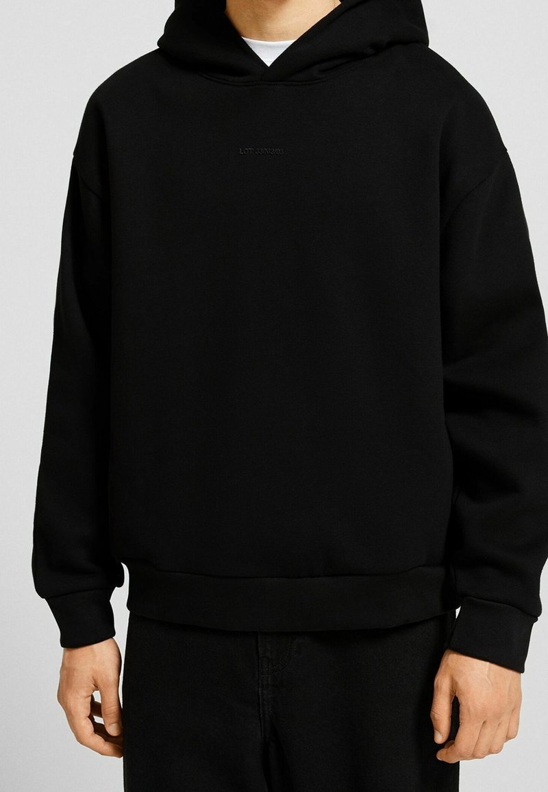 Homme portant un sweat à capuche noir uni avec un texte minimaliste sur la poitrine, associé à un pantalon noir.
