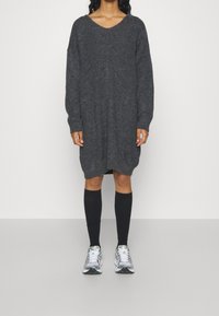 Robe pull oversize en maille gris foncé avec col en V, manches longues et fentes latérales, portée avec des chaussettes noires montantes jusqu'aux genoux et des baskets sportives.