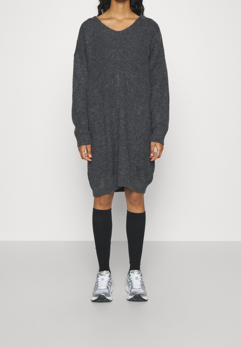 Robe pull oversize en maille gris foncé avec col en V, manches longues et fentes latérales, portée avec des chaussettes noires montantes jusqu'aux genoux et des baskets sportives.