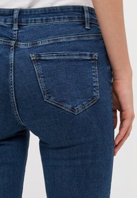 Vaqueros de mezclilla en azul oscuro con un diseño ajustado. Presenta un bolsillo trasero con una solapa triangular y costuras visibles. Textura suave en toda su superficie.