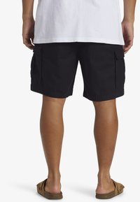 Black cargo shorts with two side pockets, made of textured fabric, knee-length, paired with brown slide sandals. Simple and practical design.