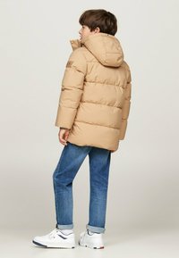 Beige pufferjack met capuchon, voorzien van een gewatteerd ontwerp, elastische manchetten en een logo-opdruk op de linkerschouder, gecombineerd met blauwe jeans en witte schoenen.