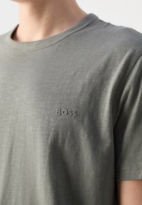 Gros plan d'une personne portant un t-shirt gris-vert à manches courtes avec "BOSS" imprimé en lettres en relief sur le côté gauche de la poitrine.