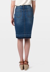 Falda de mezclilla en azul oscuro, hasta la rodilla, con bolsillos traseros, costuras decorativas y corte recto. Llevada con sandalias de cuña marrones.