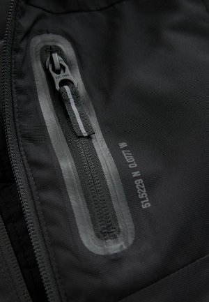 Musta ulkovaatekangas, jossa on sileä pinta, sisältää vedenpitävän vetoketjun, jonka ympärillä on heijastava nauha, sekä kokotietolapun.