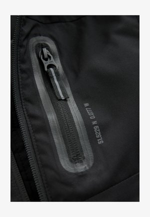 Crni vanjski materijal odjeće s glatkom teksturom ima vodootporan patentni zatvarač, obrubljen reflektirajućom trakom, i oznaku s informacijama o veličini.