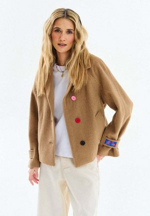 Blondynka w camelowej kurtce z różowymi, czerwonymi i czarnymi guzikami, na białej koszuli i jasnych spodniach, stojąca na białym tle.