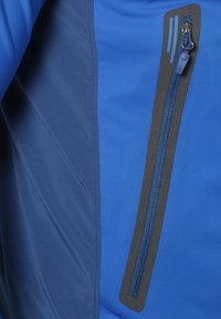 Tissu en softshell bleu avec un panneau latéral bleu foncé et une poche zippée. La fermeture éclair est dotée d'une tirette et de coutures décoratives.