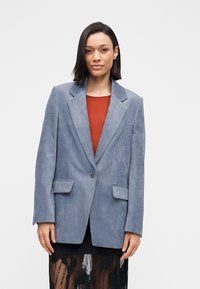 Blauwe corduroy blazer met een ingesneden revers, enkeknoopsluiting en twee voorzakken. Draagt over een rood shirt en een zwarte kanten rok.