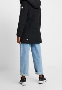 Svart pufferjacka med huva, quiltad struktur och logotyp; matchad med uppvikta ljusblå jeans och svarta sneakers.