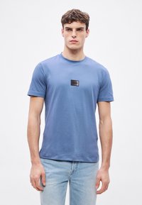 Modré bavlněné tričko s krátkými rukávy, kulatým výstřihem a černou logovou nášivkou na hrudi. Hladká textura, pohodlný střih a minimalistický design.