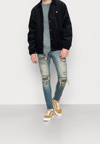 Czarna kurtka z korduroy z guzikami, szary t-shirt, zniszczone jasnoniebieskie dżinsy i beżowe trampki z białymi podeszwami.