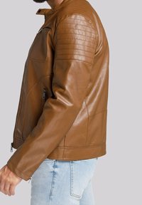 Giacca di pelle marrone con dettagli trapuntati sulle spalle, tasche con zip e vestibilità aderente, indossata sopra jeans azzurri chiari. Texture liscia.