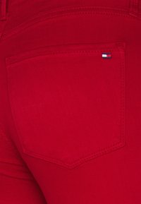 Jeans de mezclilla rojos que presentan un bolsillo trasero con un pequeño logotipo de la marca. La tela tiene una textura suave con detalles de costura doble.