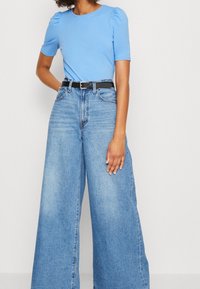 Světle modrý top s krátkým rukávem a nabranými rameny, zkombinovaný s širokými světle modrými džíny a černým opaskem se stříbrnou sponou.