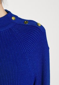 Ribbade blå tröja med hög krage, som har gyllene dekorativa knappar på axeln och ett strukturerat stickmönster.