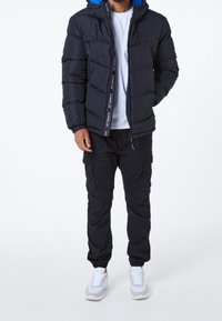 Uomo che indossa un giubbotto piumino nero con foderatura blu sul cappuccio, una t-shirt bianca, pantaloni cargo neri e sneakers bianche, in piedi su uno sfondo bianco.
