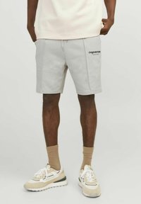 Pantalones cortos de algodón gris claro con una cintura elástica, bolsillos frontales y un logo negro en el muslo izquierdo. Combinados con zapatillas beige.