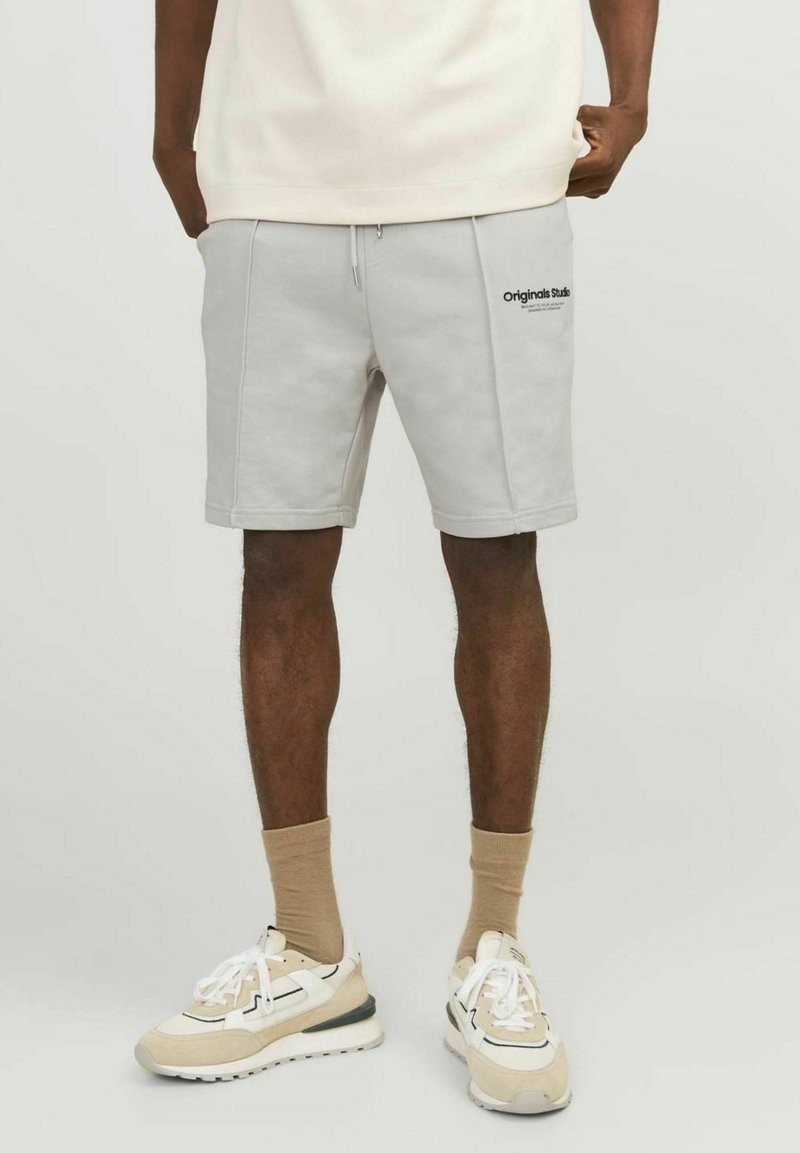 Pantalones cortos de algodón gris claro con una cintura elástica, bolsillos frontales y un logo negro en el muslo izquierdo. Combinados con zapatillas beige.