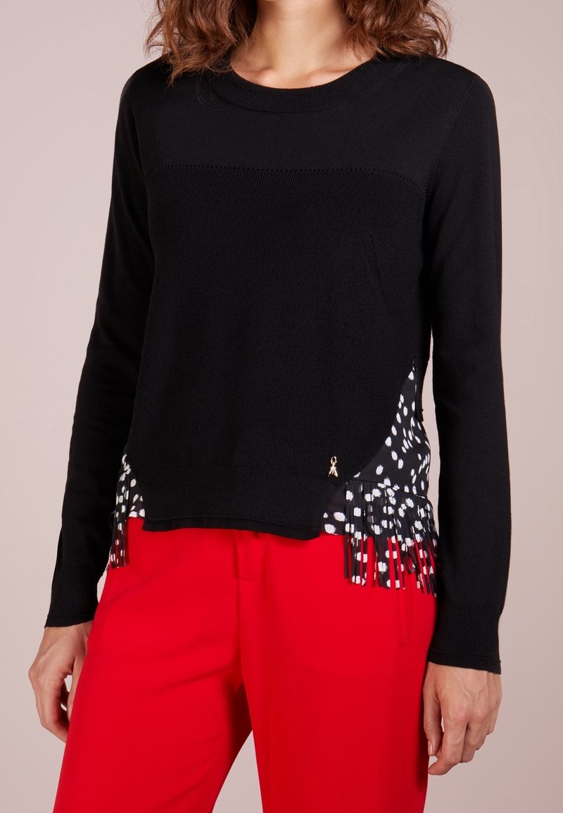 Femme portant un pull noir à manches longues avec un détail de franges tachetées noir et blanc, associé à un pantalon rouge vif.
