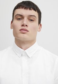 Camisa blanca con cuello y cierre de botones, hecha de una tela suave. La camisa tiene un cuello estructurado y un diseño limpio y sin adornos.