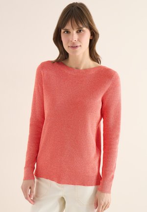 Femme aux cheveux bruns portant un pull corail à manches longues en maille et un pantalon de couleur claire, debout devant un fond uni.
