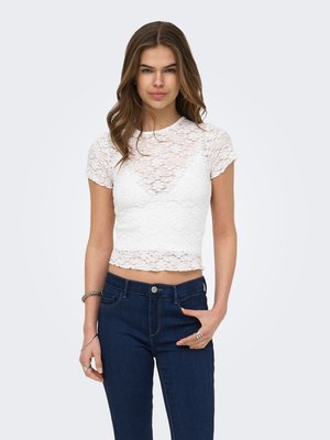 Giovane donna con lunghi capelli castani che indossa un top corto bianco in pizzo a maniche corte e jeans blu scuro, posando con una mano in tasca.