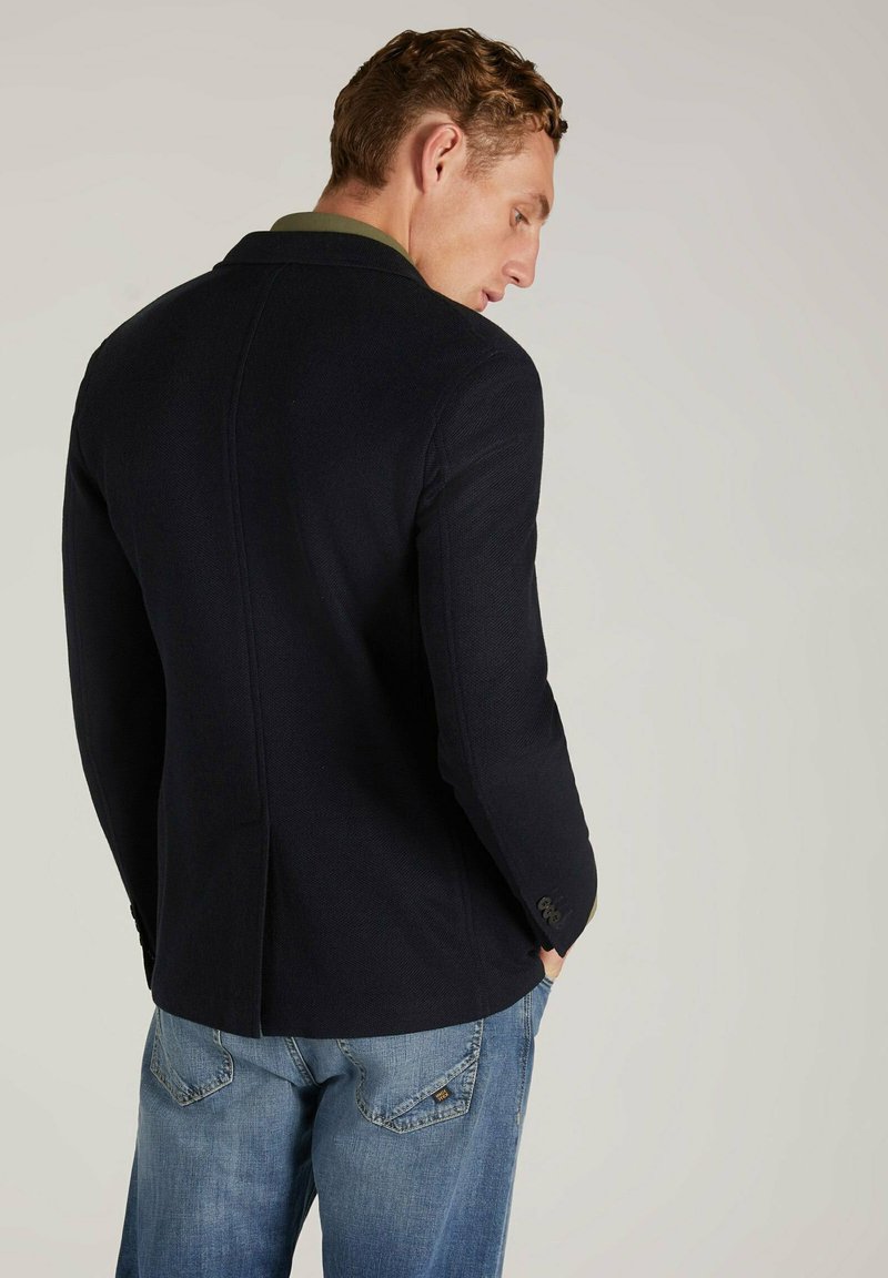 Blazer blu navy con finitura texturizzata, spacco singolo sul retro e polsini abbottonati, abbinato a jeans azzurri.