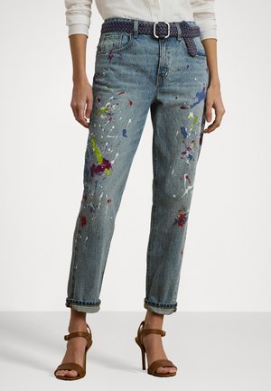 Light blue denim jeans with paint splatters in purple, green, and red. Rolled cuffs, high waist, and paired with brown strappy heels and a woven belt.