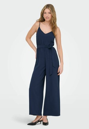 Žena oblečená v tmavomodrom bezrukávovom overal s viazaným opaskom v páse a čiernymi lodičkami na podpätku, pózujúca s jednou rukou na bok.