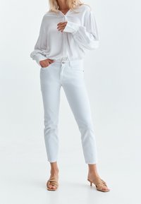 Vit skjorta med utsvängda ärmar, kombinerad med vita figursydda jeans och beigefärgade klacksandaler, mot en neutral bakgrund.